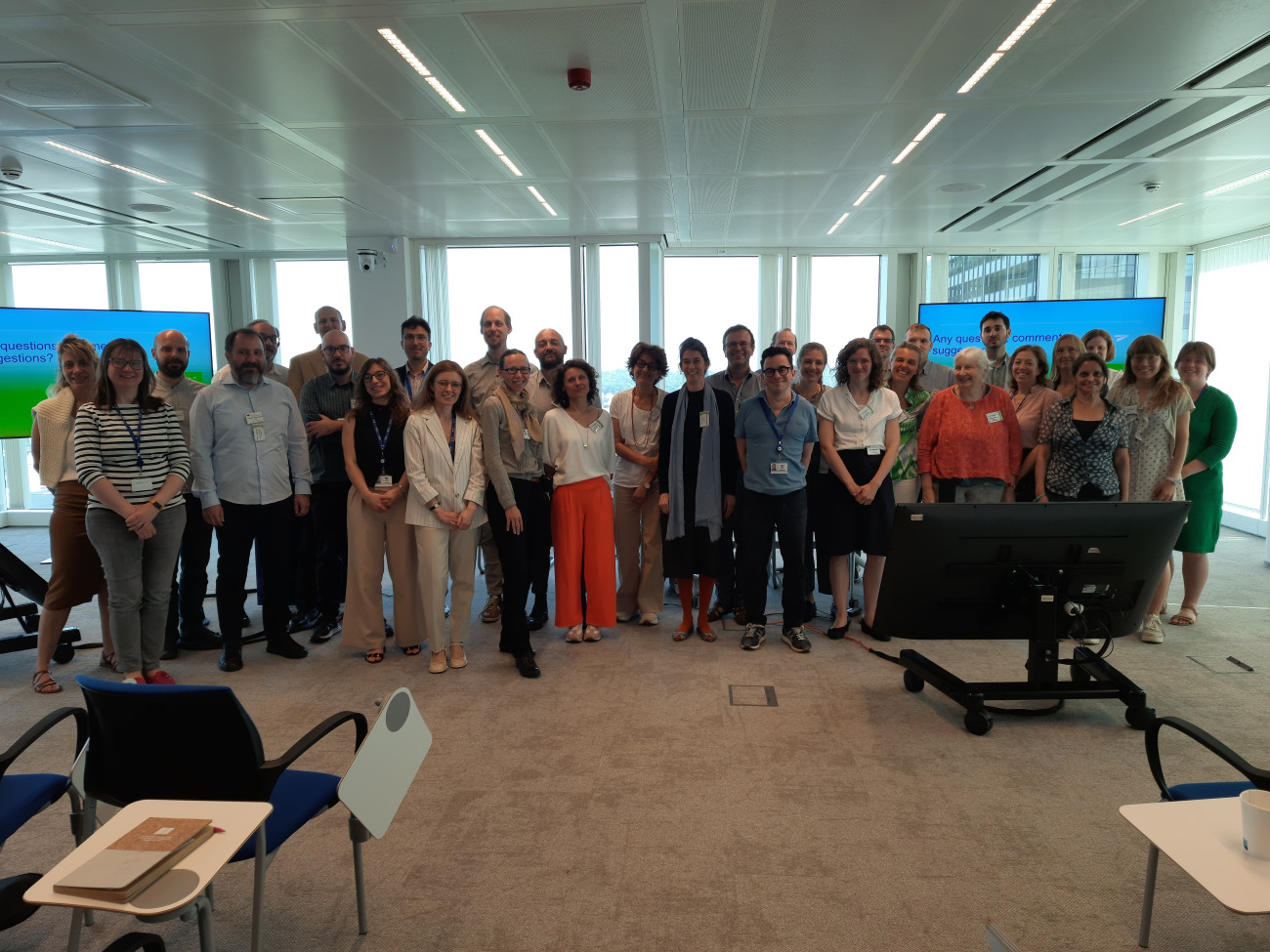 A group photo showing  a group of over 30 people  of different genders,  posing for a group picture in a conference room. Most people wear smart/casual clothes and are smiling. Behind the group, two large screens are visible, as well as several full-height windows. 