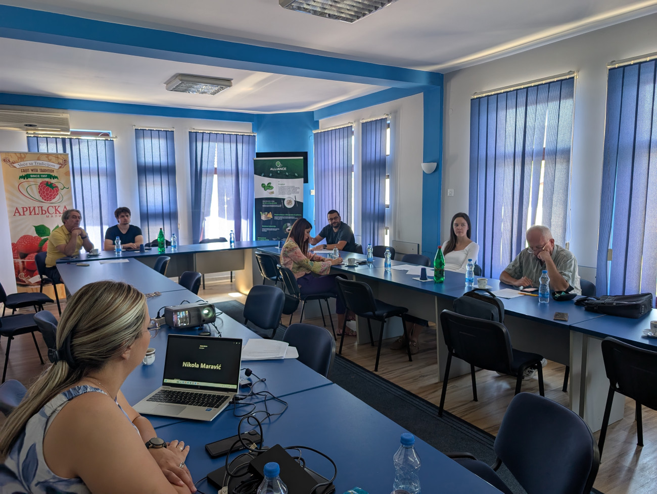 A group of people sitting around a U-shaped table. A woman has her laptop open on a  conference call with Nikola Maravic